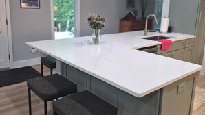 Una cocina moderna con encimera de mármol, dos taburetes de bar, un jarrón con flores y una ventana con vista a zonas verdes.