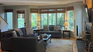 Amplia sala de estar con un sofá seccional gris grande, una mesa de centro y una chimenea. Varias ventanas ofrecen luz natural y vistas al exterior.