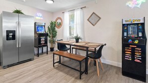 A compact kitchen with a refrigerator, a dining table with chairs, a bench, and a Pac-Man arcade machine.