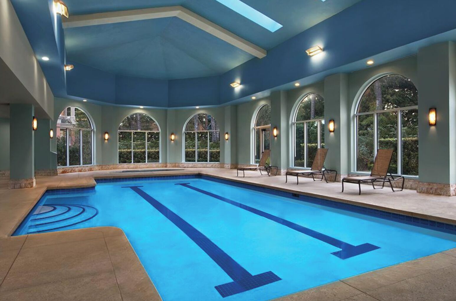 Una amplia piscina interior con marcas claras para carriles, rodeada de sillones de descanso y grandes ventanales arqueados.