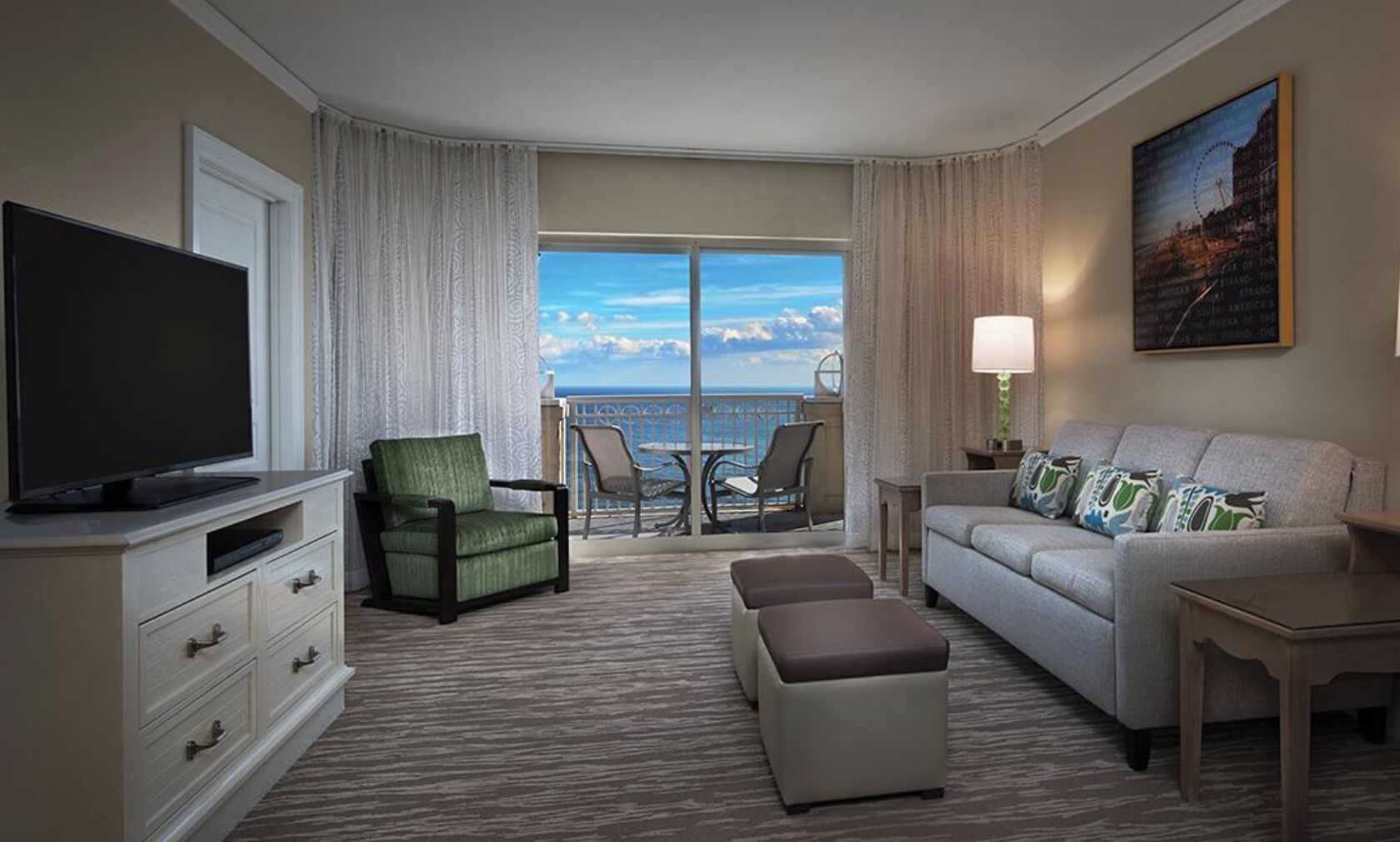 A hotel room with a large flat-screen TV, a green armchair, a white sofa, a small table, and a balcony with a view of the ocean.