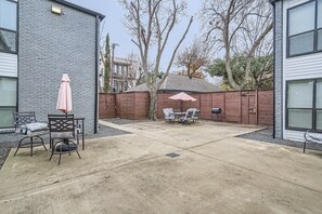 Un patio trasero con piso de concreto, dos sillas, una mesa y un paraguas rosa.
