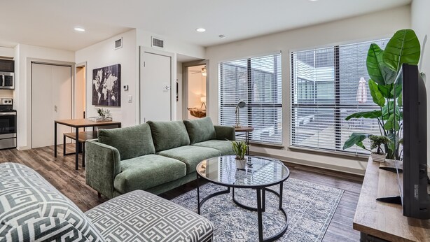 A modern living room with a green sofa, a patterned ottoman, a glass coffee table, and a view through large windows with blinds.