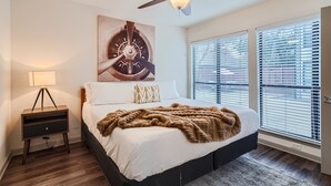 A bedroom with a bed, a ceiling fan, a nightstand, a lamp, and a window with blinds.