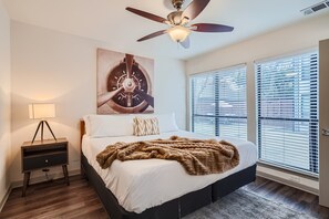 Un dormitorio con cama, ventilador de techo, mesita de noche, lámpara y ventana con persianas.