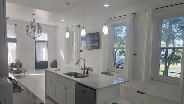 Una cocina moderna con una isla central, pileta doble y lavavajillas integrado. Hay luces colgantes, un candelabro y amplias ventanas con vistas al exterior.