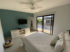 Une chambre avec un lit, une télévision, un ventilateur de plafond, une porte-fenêtre coulissante donnant sur un balcon et un petit coin salon avec un panier et un coussin.