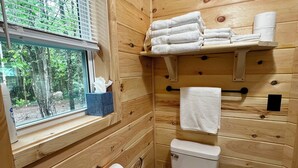 Un baño de madera con inodoro, un estante con toallas, una ventana y un rollo de papel higiénico.