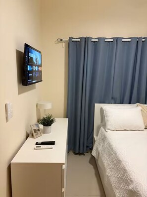 Une chambre d’hôtel avec un lit, un bureau doté d’un écran d’ordinateur, une plante et un cadre photo.