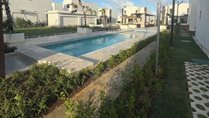A modern pool area with a clear blue sky, surrounded by white buildings and greenery.