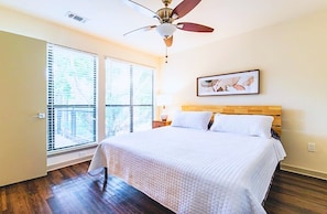 Un dormitorio con cama, ventilador de techo, ventana con persianas y un cuadro en la pared.