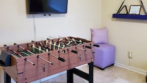 Quarto com mesa de pebolim, televisão fixada na parede, um banquinho roxo e uma prateleira com uma placa de boas-vindas.