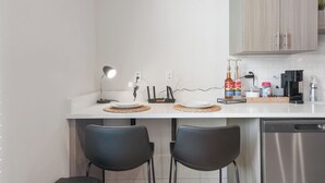 Eine moderne Küche mit weißen Arbeitsplatten, einer Kaffeemaschine, einem Toaster und einer Saucenflasche. Zwei schwarze Stühle und ein weißer Tisch sind ebenfalls vorhanden.