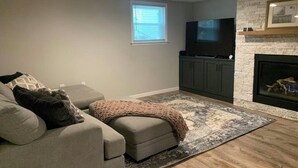 A living room with a gray sofa, ottoman, and a TV on a stand. - We Are…..2 miles from Beaver Stadium! (State College)