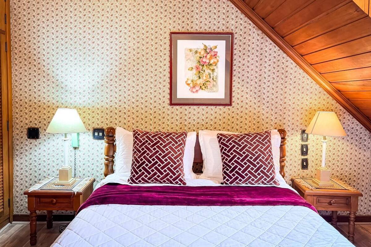 A bedroom with a sloped ceiling, a bed with patterned pillows, a framed floral artwork on the wall, and bedside lamps.