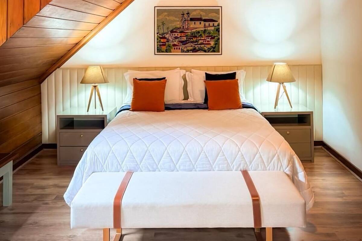 A bedroom with a sloped ceiling, a bed with a quilted cover, two bedside lamps, and a painting on the wall.