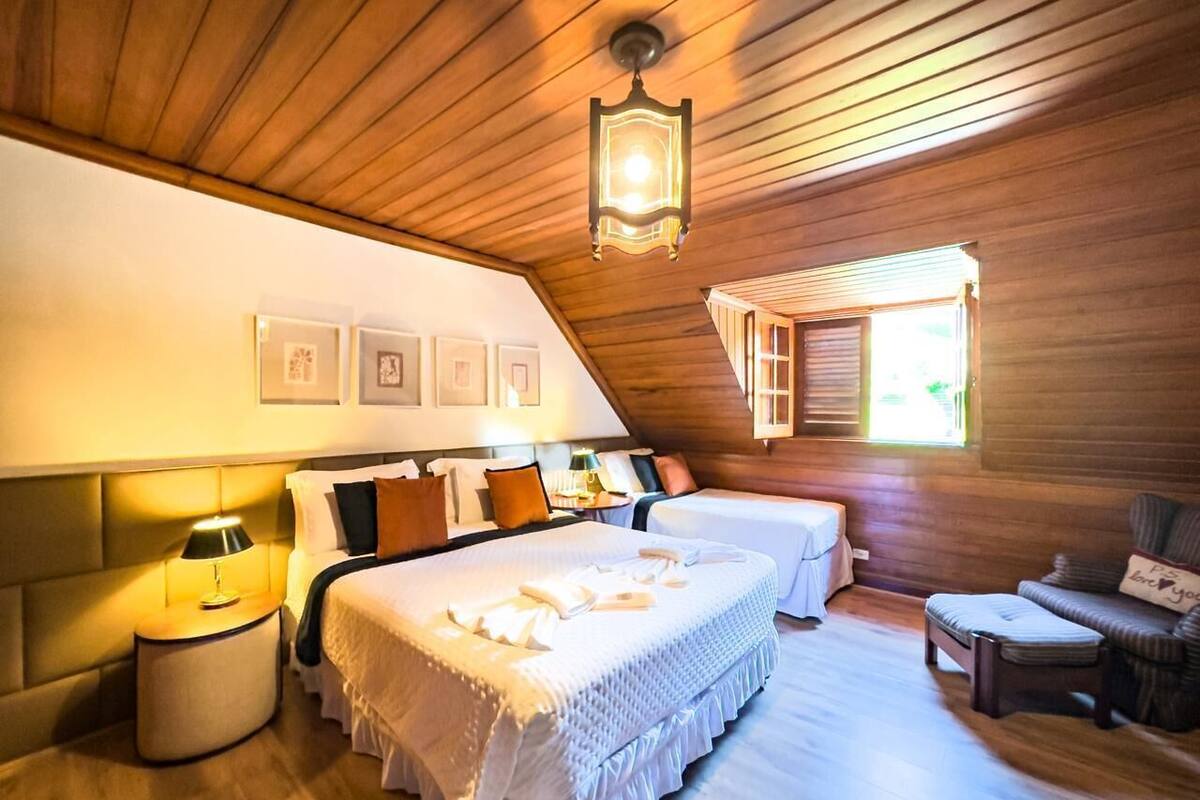 A cozy bedroom with a large bed, a smaller bed, a nightstand, a lamp, a chair, and a window with shutters.