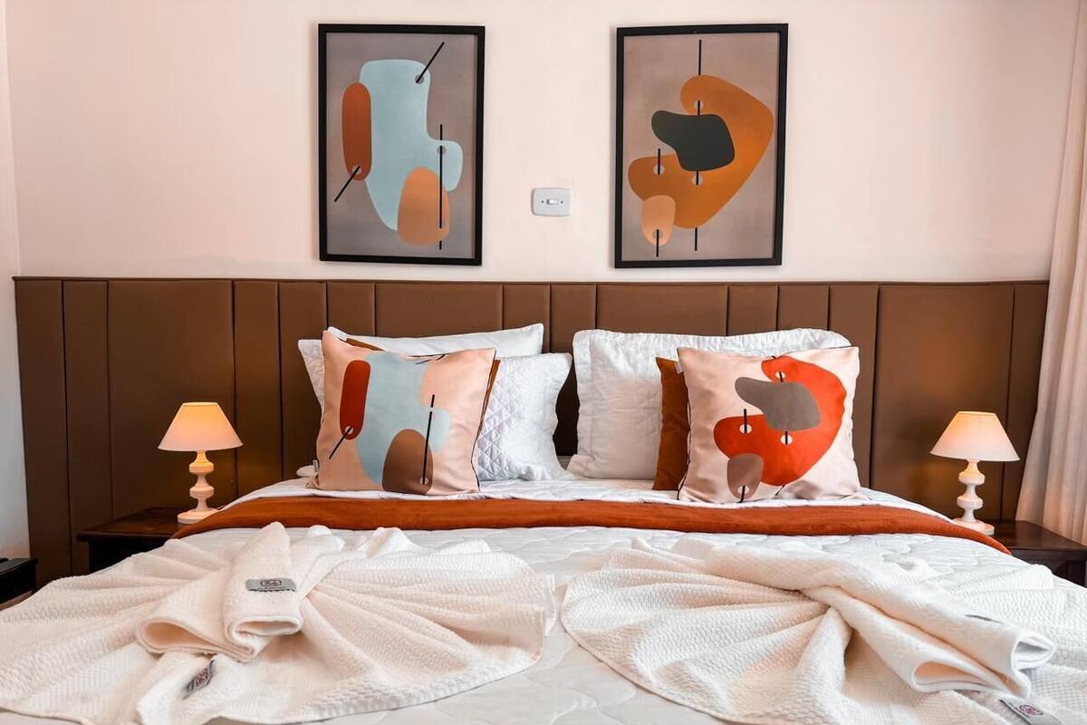 A hotel room with a bed, pillows, a headboard, two wall-mounted lamps, and abstract art on the wall.