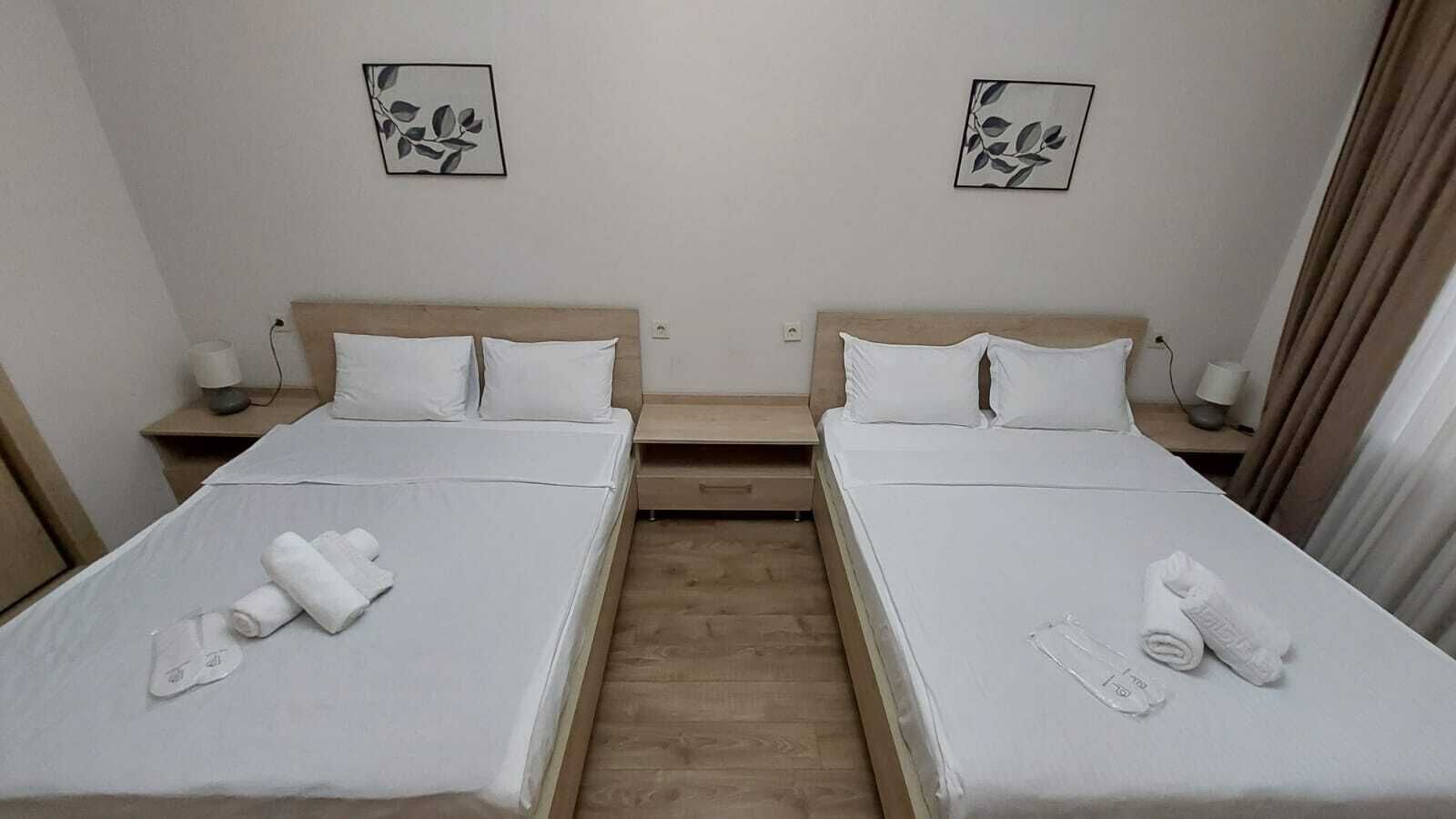 A hotel room with two beds, each with white linens and towels. There are two bedside tables with lamps, and the walls are adorned with leaf-patterned artwork.