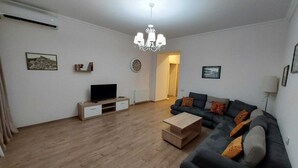 Una sala de estar con un sofá de color gris oscuro, una mesa de centro de madera, un televisor sobre un mueble de madera y una araña.