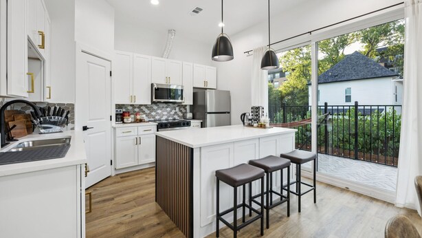 Une cuisine moderne avec des meubles blancs, un îlot central, des tabourets de bar et une vue sur un quartier résidentiel.