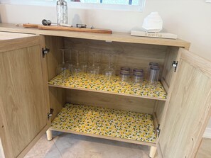 A wooden cabinet with three shelves, each holding wine glasses and jars.