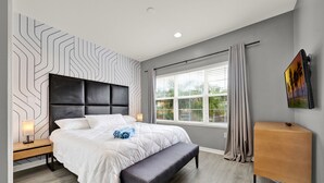 Un dormitorio moderno con una cama grande, un banco, una mesita de noche, una ventana con cortinas y un televisor montado en la pared.