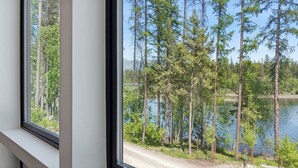 A view through a window of a forested area with a lake and a dirt path. - Newly built two bedroom rental with Blanchard Lake access in Whitefish! (Whitefish)