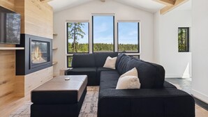 A modern living room with a black sectional sofa, a wooden coffee table, and a fireplace. Large windows offer views of a forested landscape. - Newly built two bedroom rental with Blanchard Lake access in Whitefish! (Whitefish)