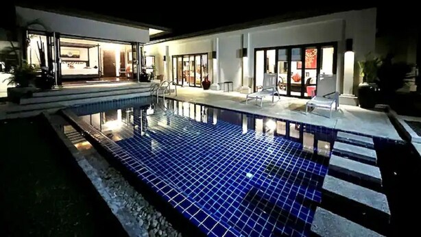 A swimming pool with a tiled deck, a covered seating area, and a building with large glass doors.