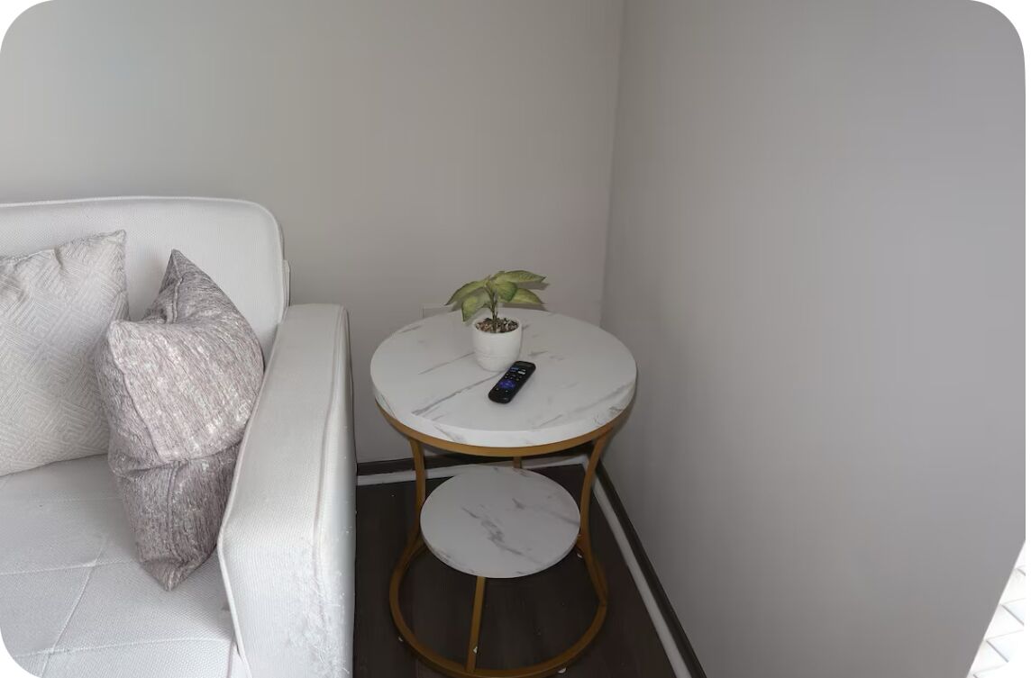 Una mesita pequeña y redonda, de color blanco, con una planta en maceta y un control remoto, ubicada junto a un sofá blanco con cojines estampados.