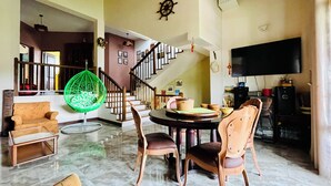Amplia sala de estar con mesa de comedor, sillas, una butaca colgante verde, una escalera y un televisor de pantalla plana.