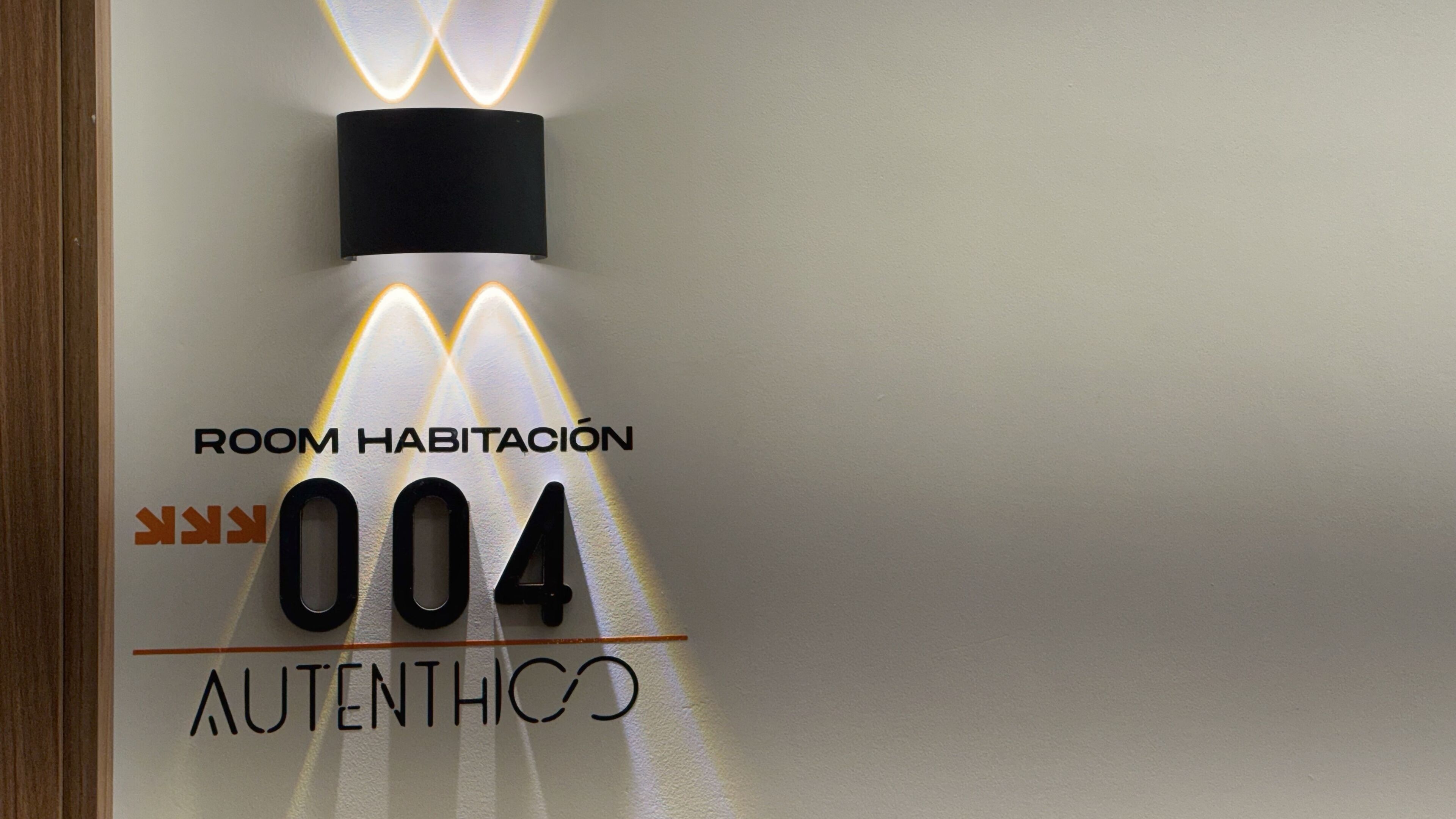 Señal en la pared con la leyenda 'ROOM HABITACIÓN 004' y 'AUTÉNTICO', indicando el número y nombre de la habitación.