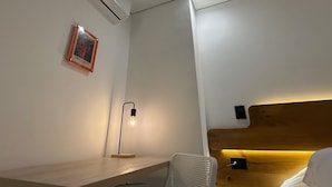 A hotel room with a wooden desk, a wall-mounted air conditioner, a framed picture, a desk lamp, and a bed with a wooden headboard.