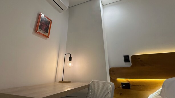 A hotel room with a wooden desk, a wall-mounted air conditioner, a framed picture, a desk lamp, and a bed with a wooden headboard. - Autenthico Local Stay Hotel Envigado (Envigado)