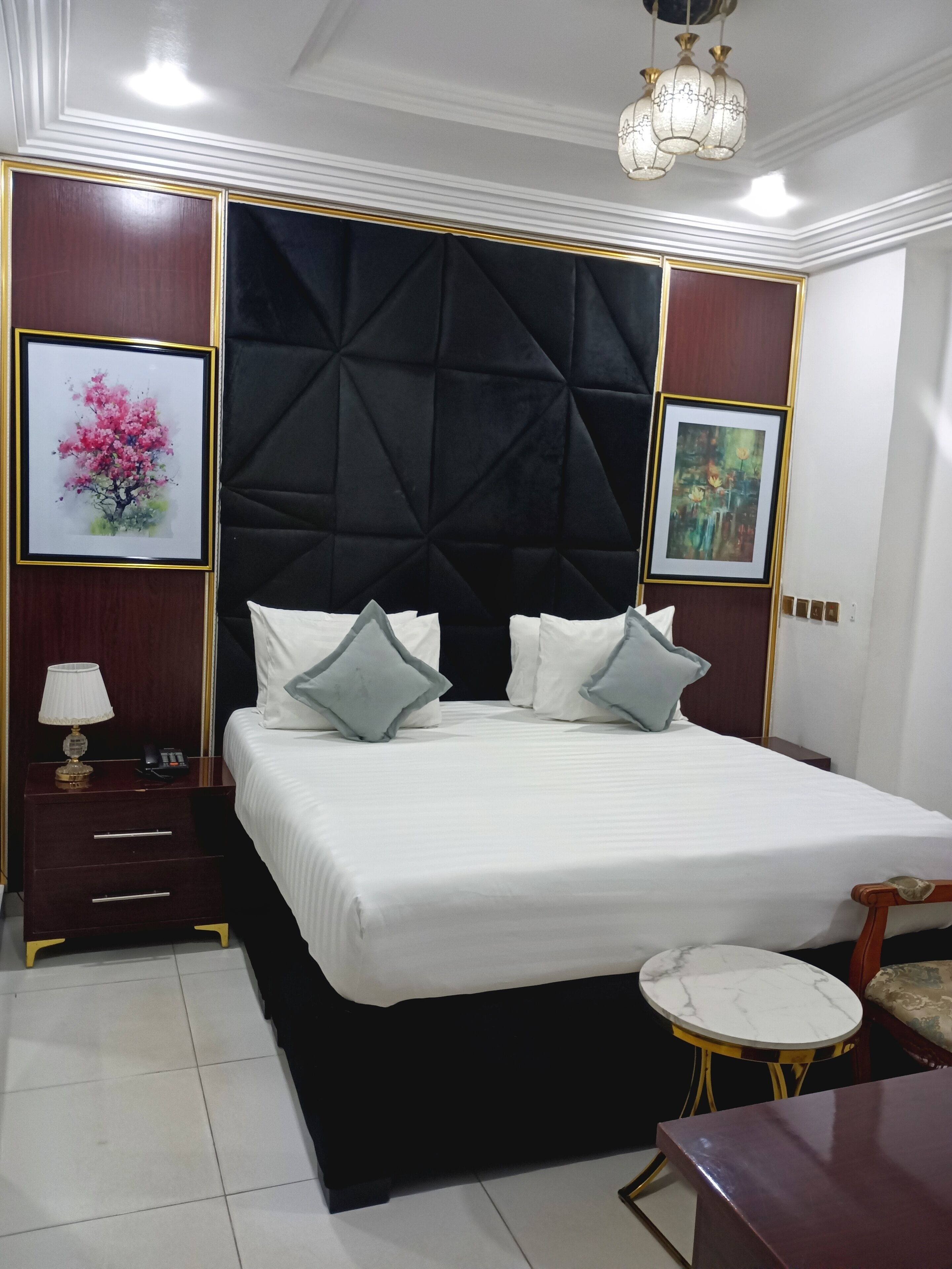A hotel room with a large bed, a nightstand with a lamp, a small round table, and two framed artworks on the walls.