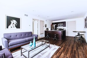 Una sala de estar moderna con un sofá gris, una mesa de centro de vidrio y una cocina con armarios oscuros y un microondas.