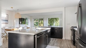 Une cuisine moderne dotée d’un grand îlot central, de meubles noirs, d’appareils électroménagers en acier inoxydable et de grandes fenêtres offrant une vue sur la verdure.