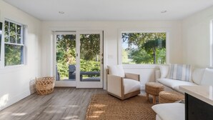 Un salon moderne comprenant un canapé, un fauteuil et un pouf, de grandes fenêtres donnant sur un paysage verdoyant, et une porte-fenêtre coulissante qui mène à l’extérieur.