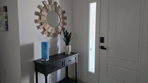 A hallway with a white door, a black console table with a blue vase and a potted plant, and a decorative mirror with a wooden frame. - Beautiful, well appointed, 3-bed house in DeLuna - 2 Kings! (The Villages)