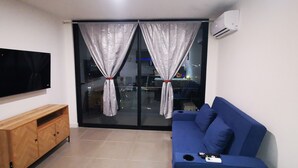 A living room with a TV, a blue sofa, a wooden cabinet, and a sliding glass door with curtains. - Luxury apartment full equip. in San Jose with balcony city views & top amenities (San José)