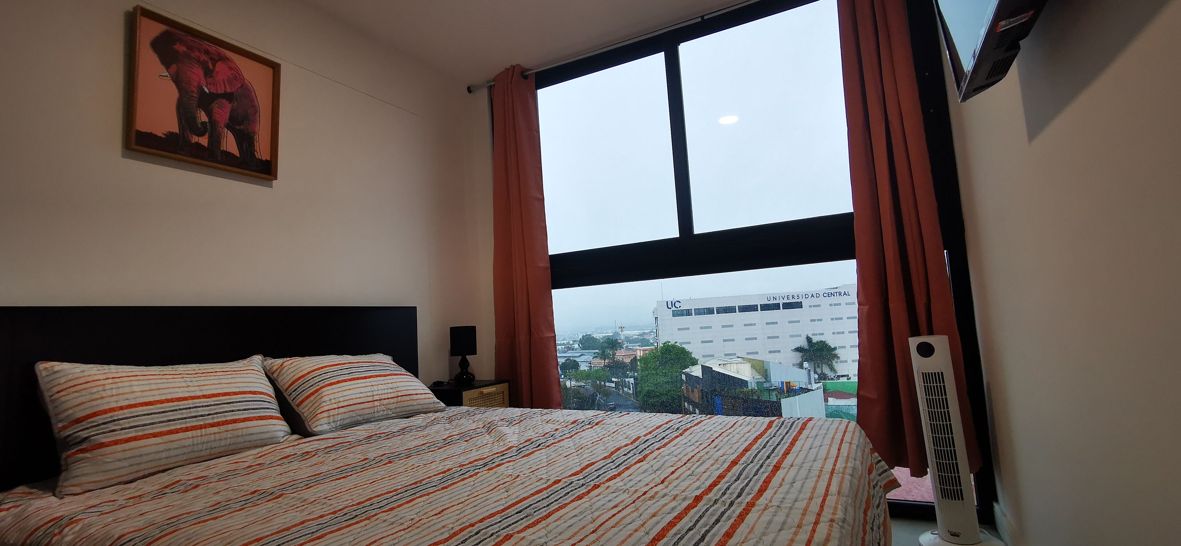 Habitación de hotel con una cama grande, una mesita de noche, una lámpara, una ventana con cortinas y vistas a un edificio.