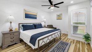 A bedroom with a bed, nightstands, a ceiling fan, a window with blinds, and a painting on the wall.