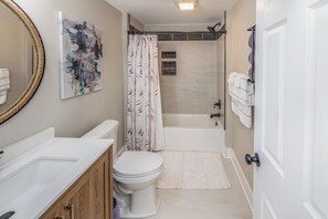 Une salle de bain moderne avec un lavabo, des toilettes et une baignoire blancs. On y trouve un rideau de douche à motifs floraux et un grand miroir avec un cadre en bois.