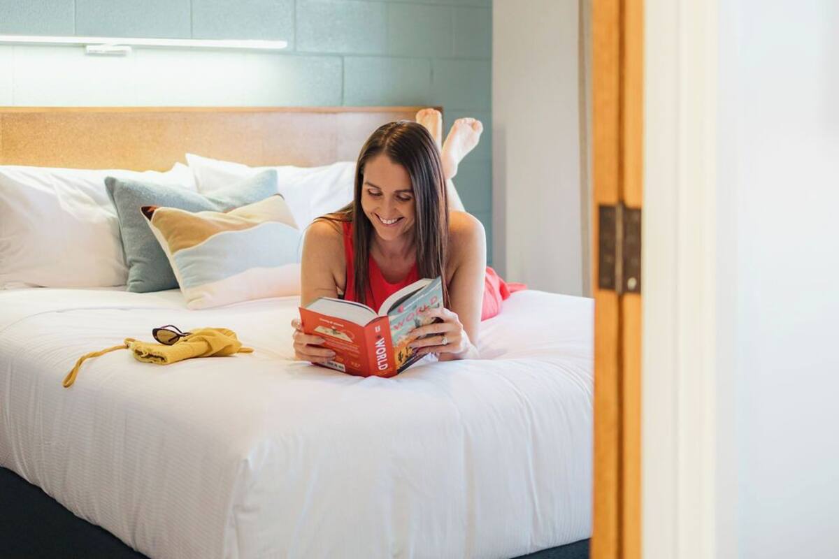 A person reading a book on a bed with a blue headboard, a yellow towel, and a pair of sunglasses on the bed.