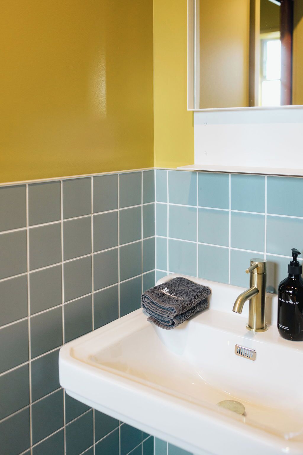 A bathroom with a white sink, a gold faucet, and a mirror above it. There are blue and gray tiles on the walls.