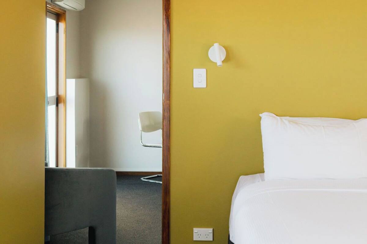 A hotel room with a bed, a chair, and a desk. There is a door leading to another room.