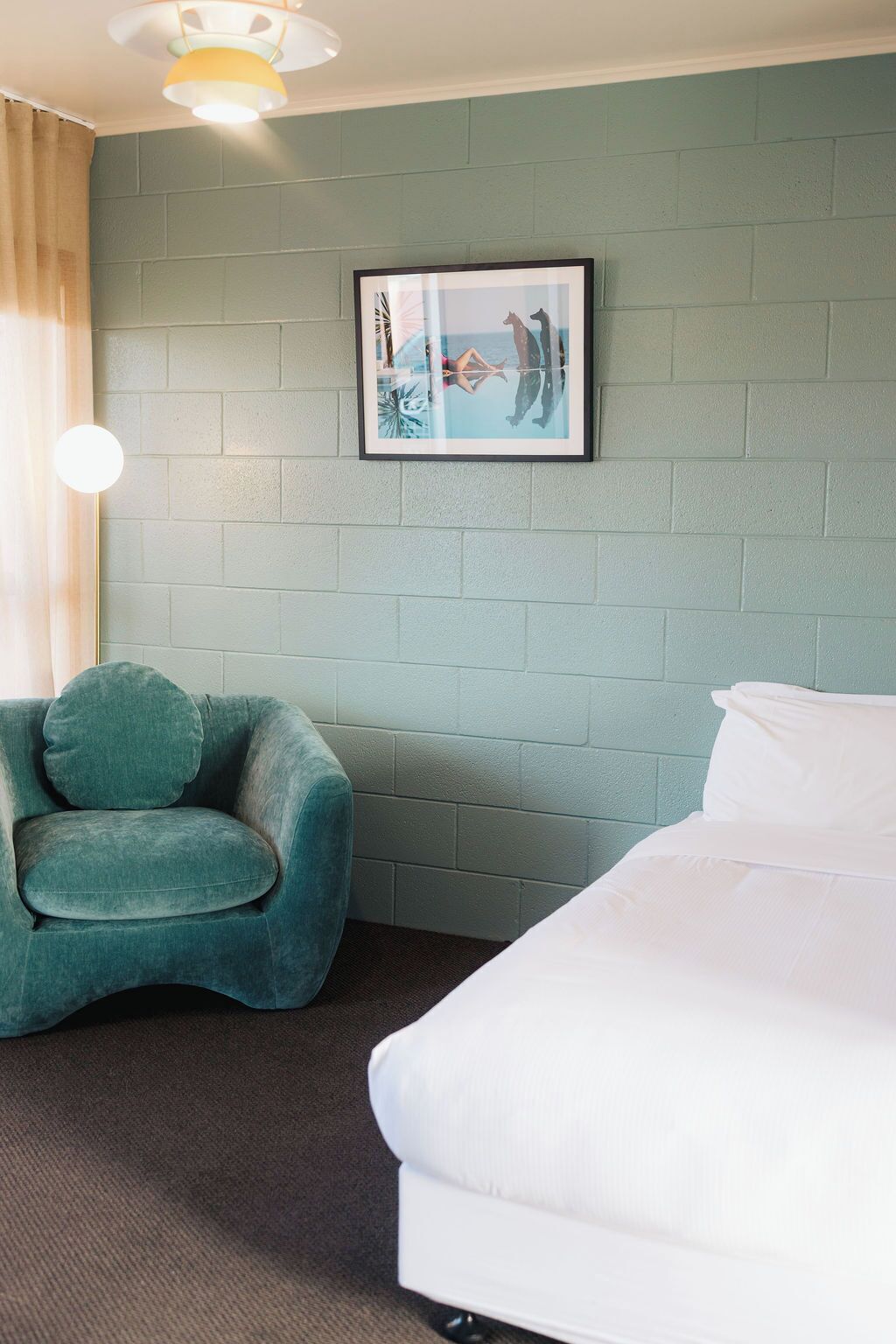 Une pièce avec un mur en briques de couleur turquoise, un tableau encadré, un fauteuil turquoise et un lit blanc.