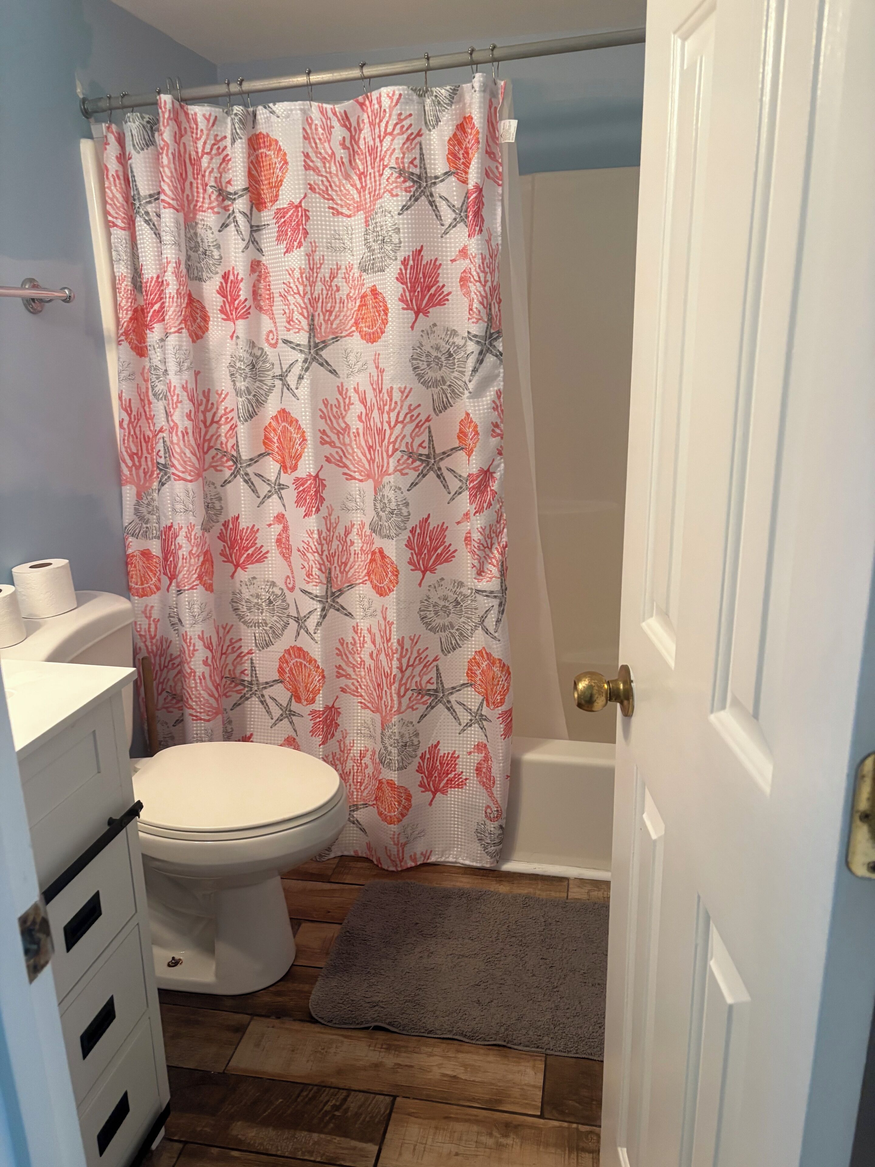 Une salle de bain avec des toilettes, un rideau de douche orné d’un motif corail et d’étoiles de mer, un plancher en bois et une porte blanche.