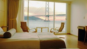 A hotel room with a bed, two chairs, a desk, and a large window with a view of a bridge and sunset. - HOTEL SETRE KOBE MAIKO (Kobe)